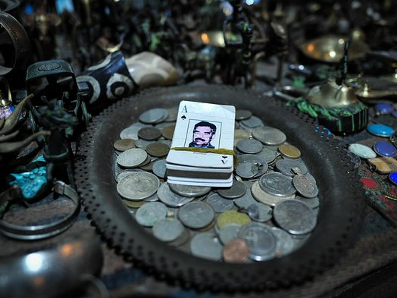 A deck of cards with Saddam Hussein on it sits on a plate full of coins, which is on a table of trinkets