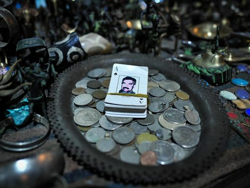 A deck of cards with Saddam Hussein on it sits on a plate full of coins, which is on a table of trinkets