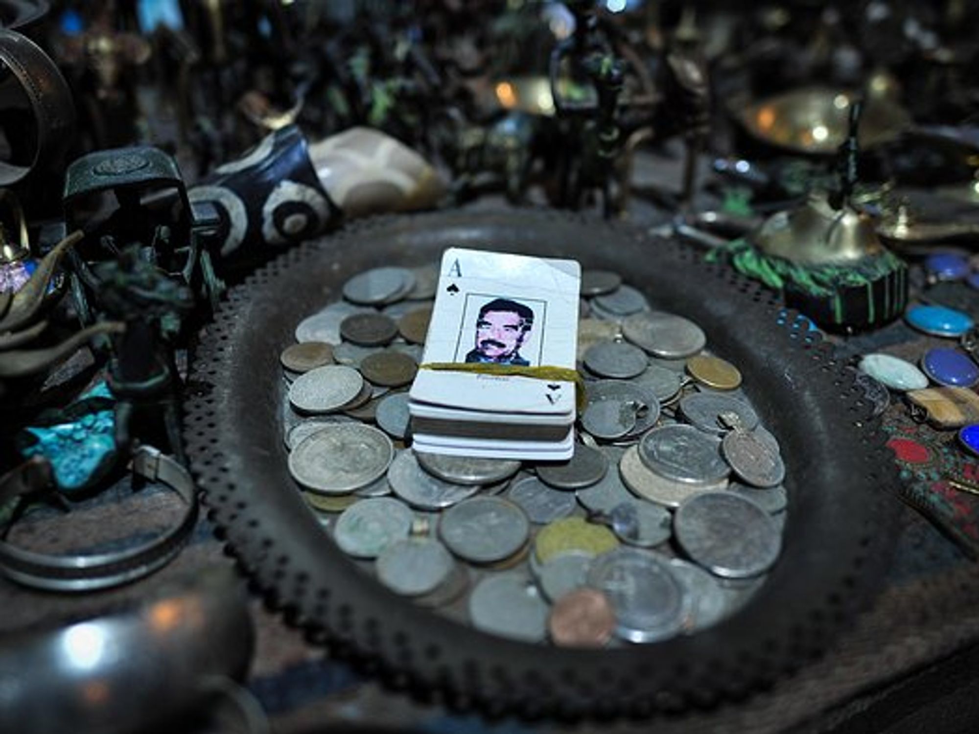 A deck of cards with Saddam Hussein on it sits on a plate full of coins, which is on a table of trinkets
