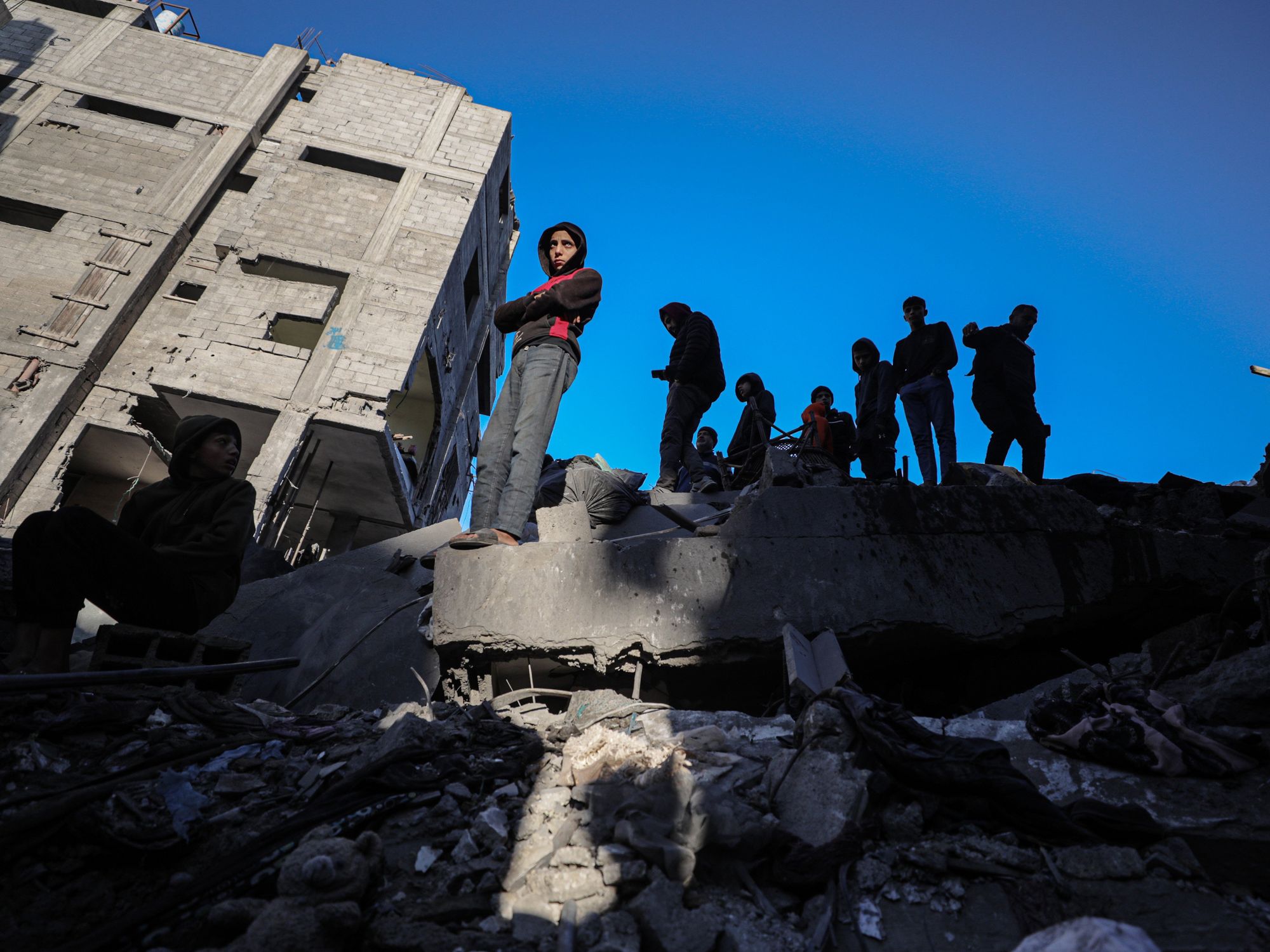 A group of people mostly young boys, stand on a pile of rubble as one of them is illuminated by the sun and the rest are in the shadows.
