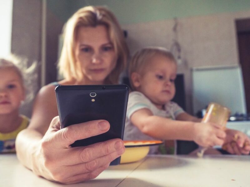 A mother holds her two toddlers as she looks into her cellphone.
