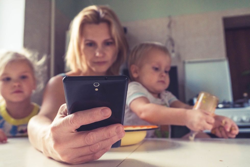 A mother holds her two toddlers as she looks into her cellphone.