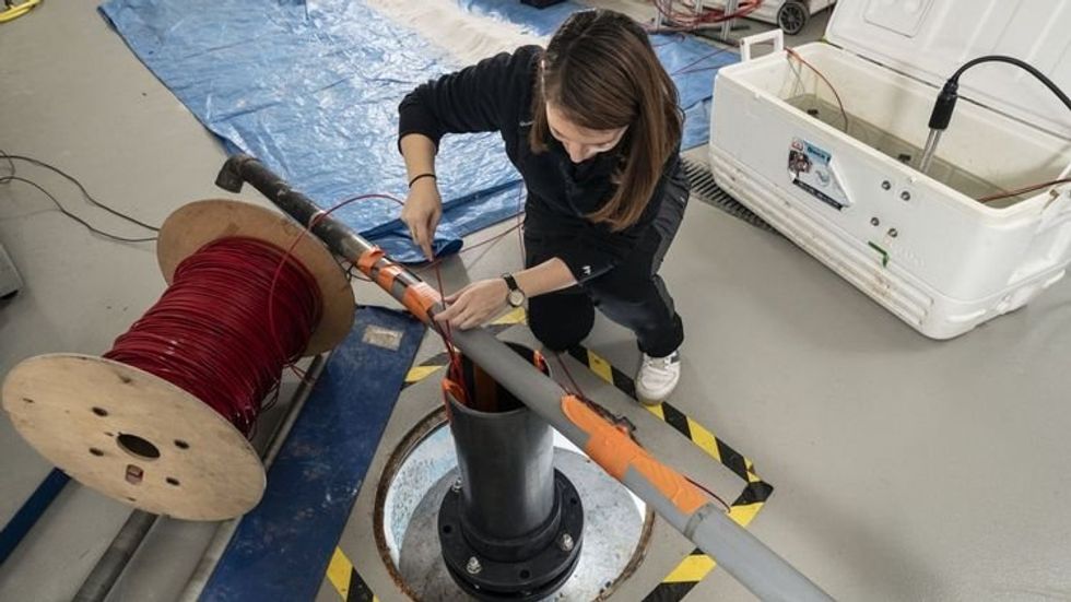 A researcher kneels in front of a pipe going into the ground, appearing to feed a wire down into the pipe