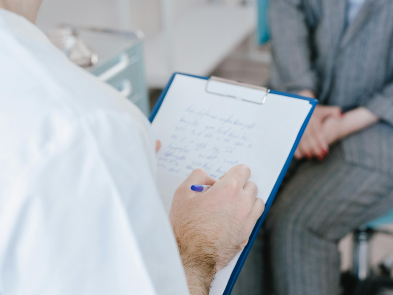 Photo of a male doctor writing on a notepad, with a female patient in the background