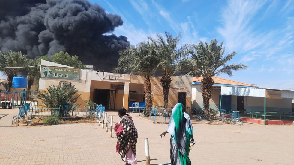 Photo of people walking by a building in Khartoum with smoke rising in the background after a bombing at a fuel station.