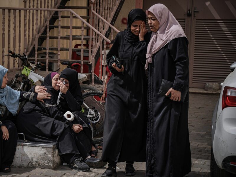 5 women in hijabs gathered together to mourn, 3 sitting, 2 standing