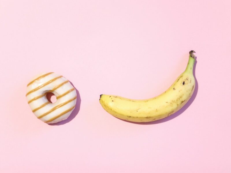 A banana and donut sit with a pink background.