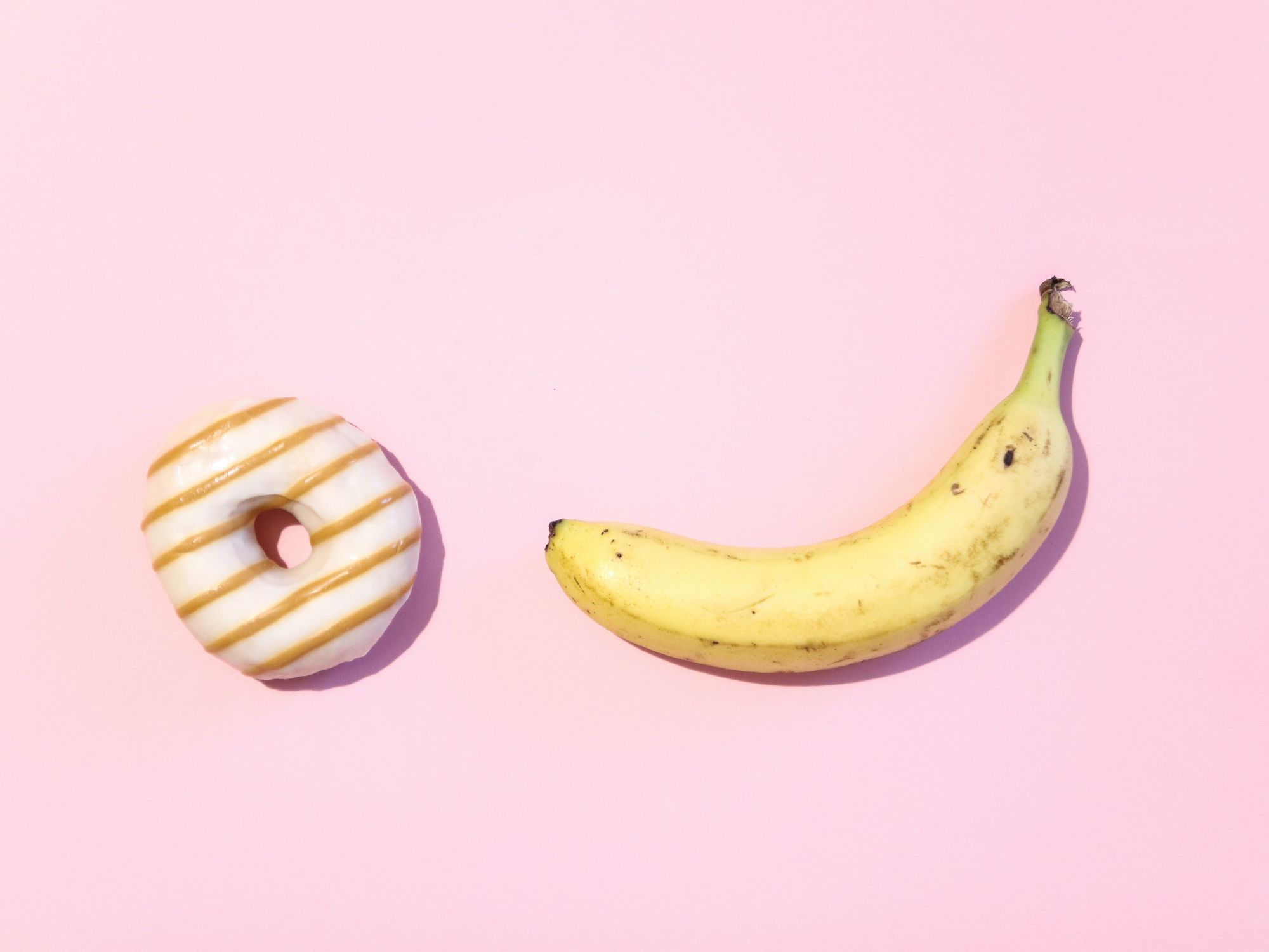 A banana and donut sit with a pink background.