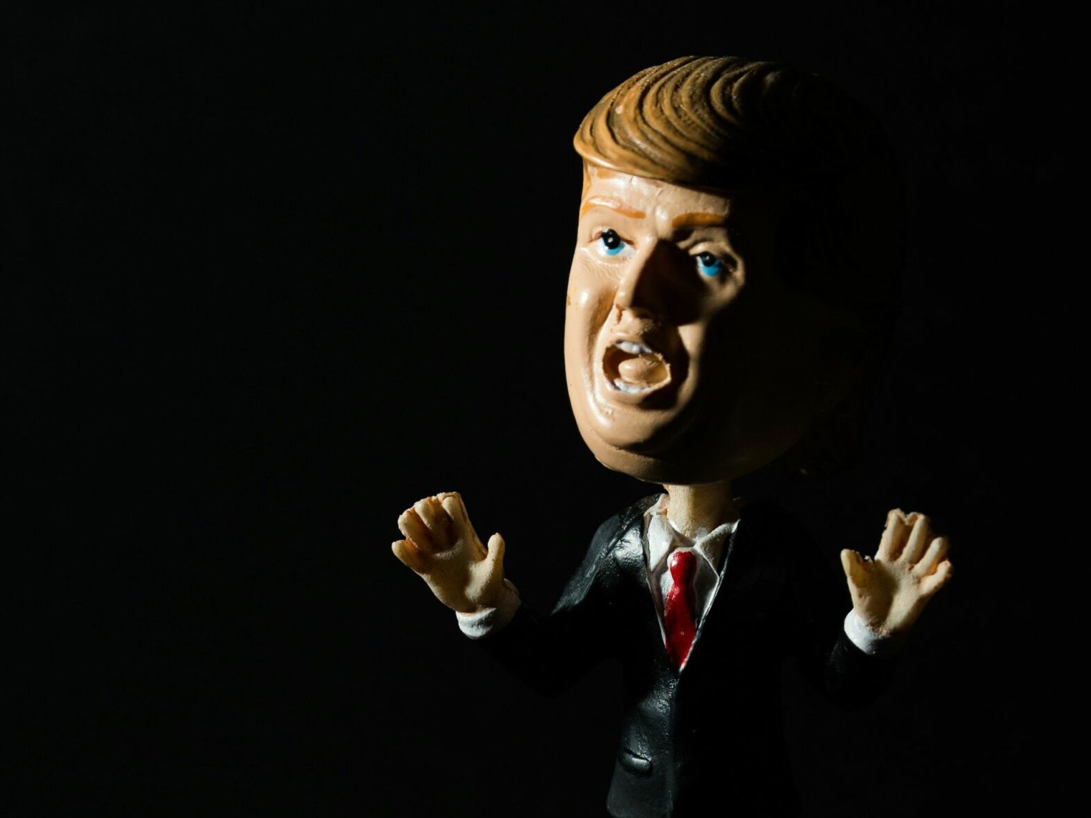 A bobblehead of Donald Trump is lit from one side, with a shadow on the other in a very dark setting