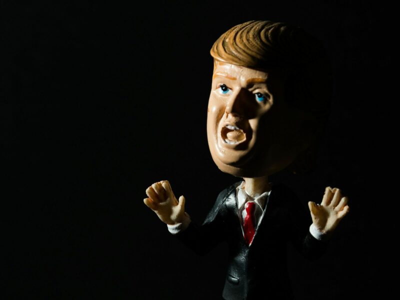 A bobblehead of Donald Trump is lit from one side, with a shadow on the other in a very dark setting