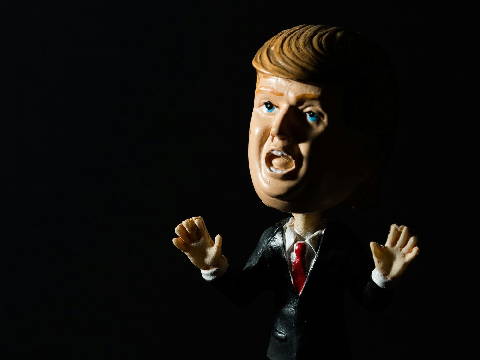 A bobblehead of Donald Trump is lit from one side, with a shadow on the other in a very dark setting