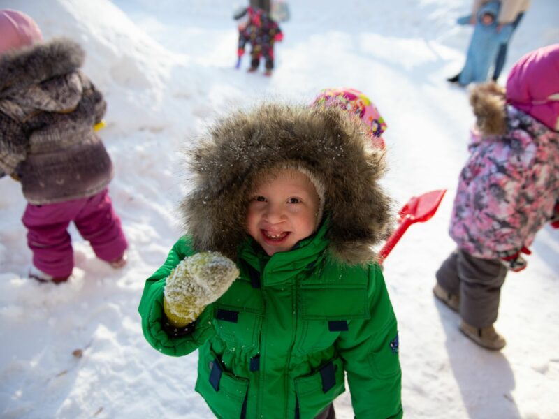 A child in a green coat is seen in the foreground of a snowy landscape, with other children seen behind them