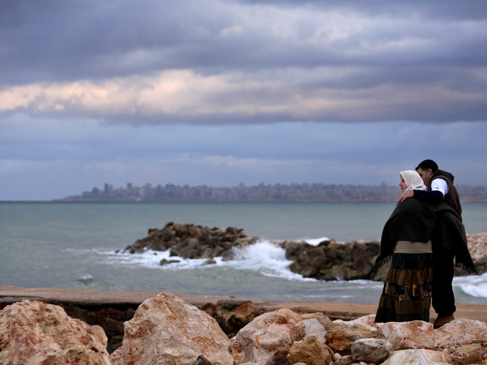 A couple looking at the sea at sunset. In the distance, the city of Beirut. Sidon, Lebanon.