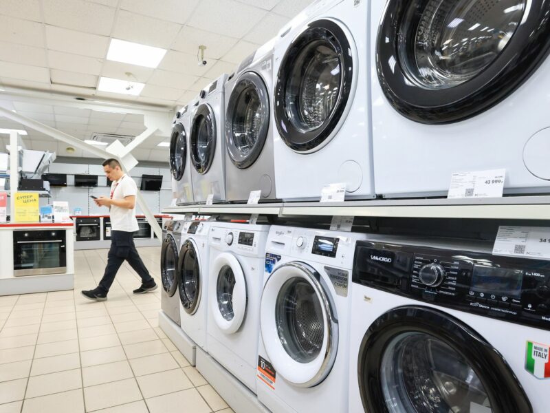 A double stack row of washing machines for sale with price tags. A man is walking in the background on his phone.