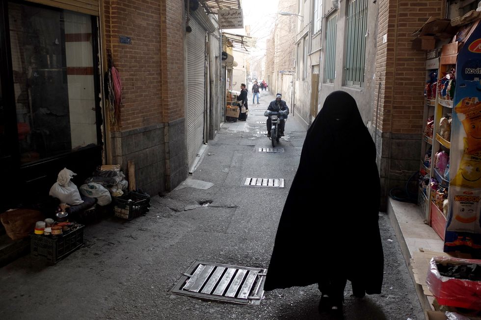 A fully covered woman walks in a dark alleyway as a man on a motorcycle is seen in the distance