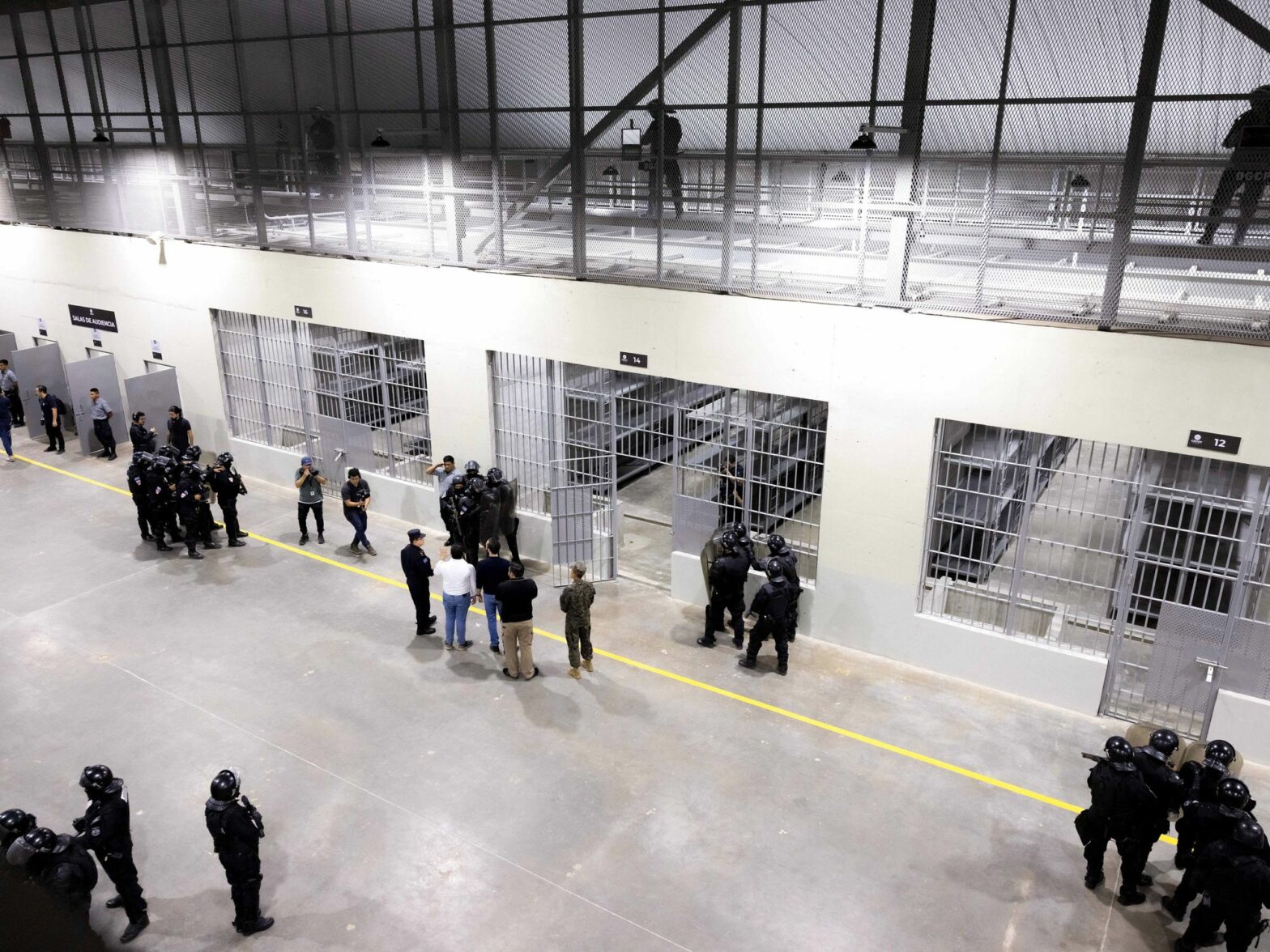 A large open prison is seen from a catwalk overlooking a main central area with barred-in sections. Men in riot gear are clumped around the room.
