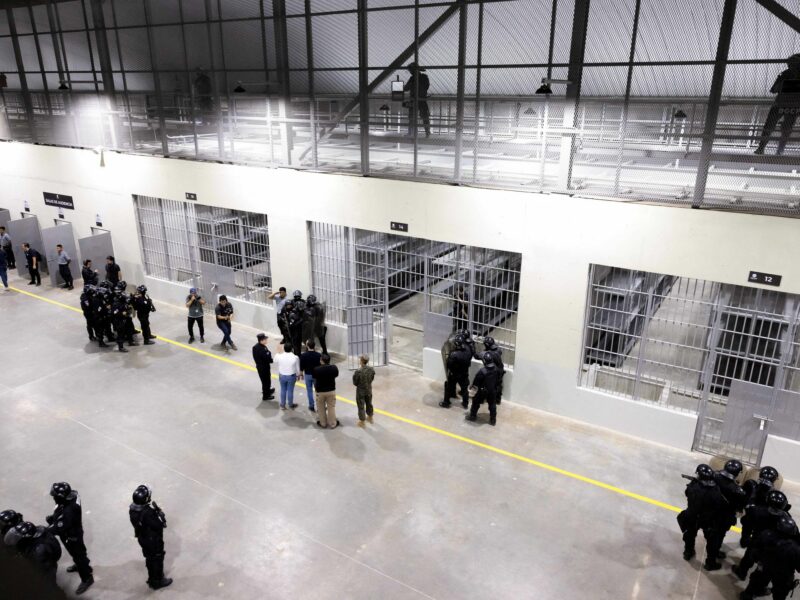 A large open prison is seen from a catwalk overlooking a main central area with barred-in sections. Men in riot gear are clumped around the room.