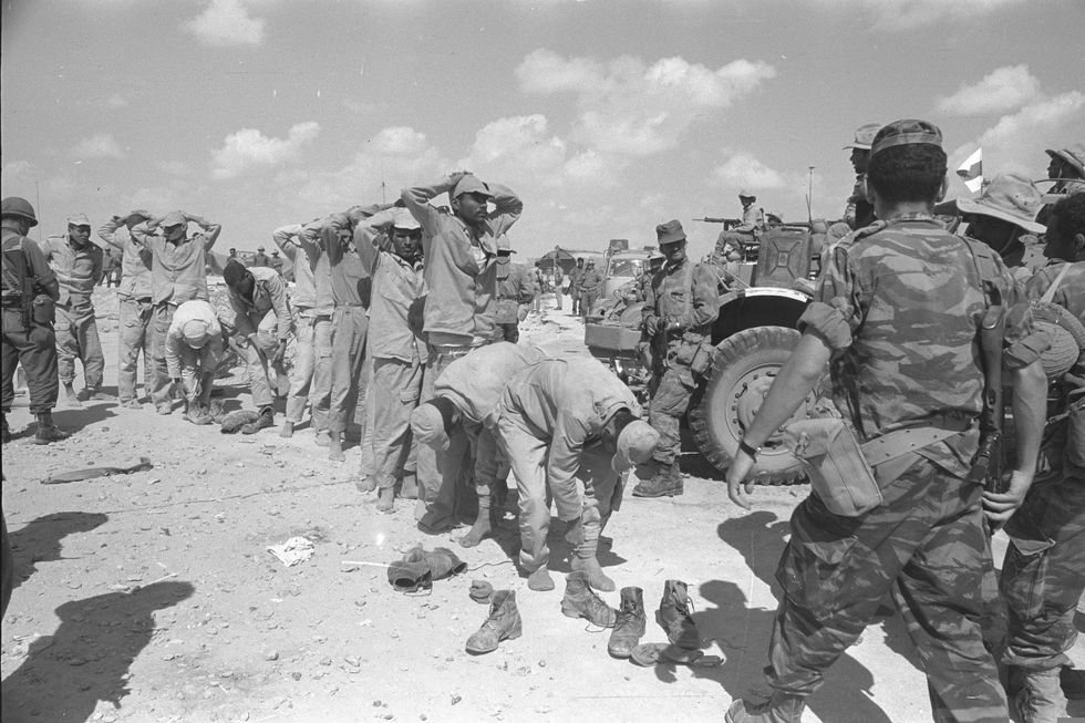 A line of people are surrounded by soldiers as some take their shoes off and others keep their hands on their head.