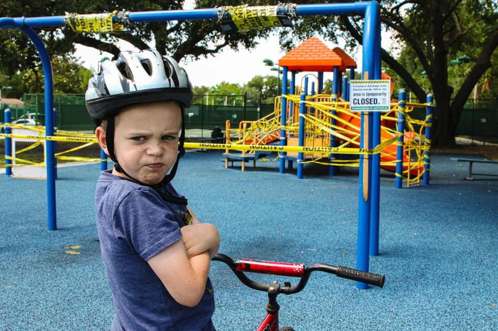 A little boy looks off into the distance angrily, with his arms crossed over his chest. He is angry because the playground is closed.