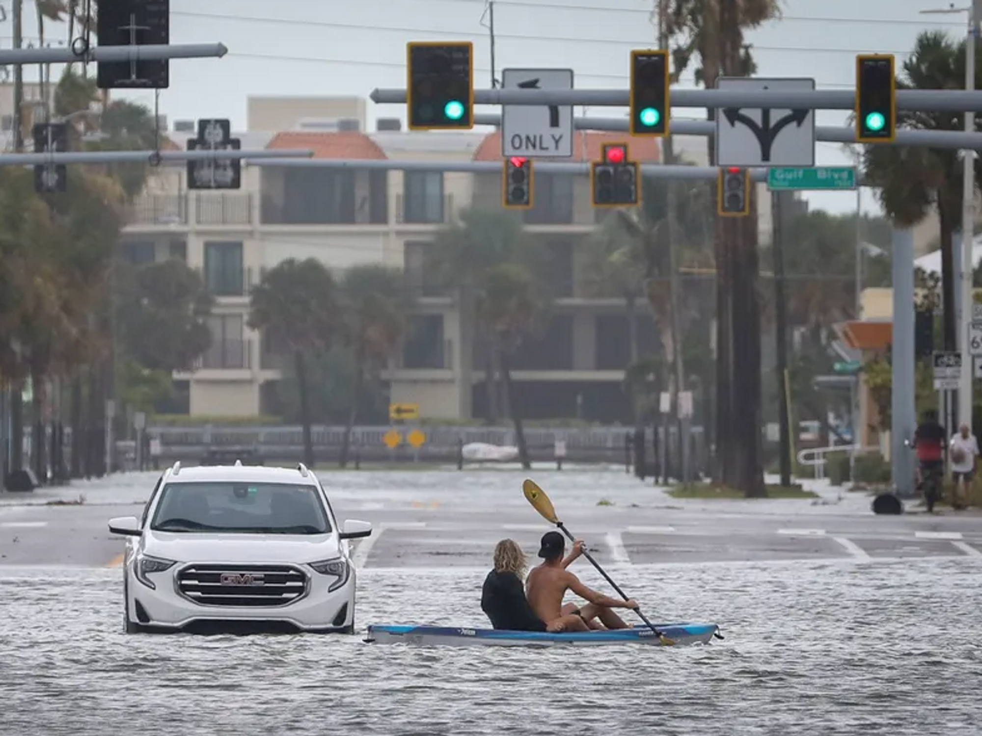 A man and woman kayak past an abandoned vehicle.