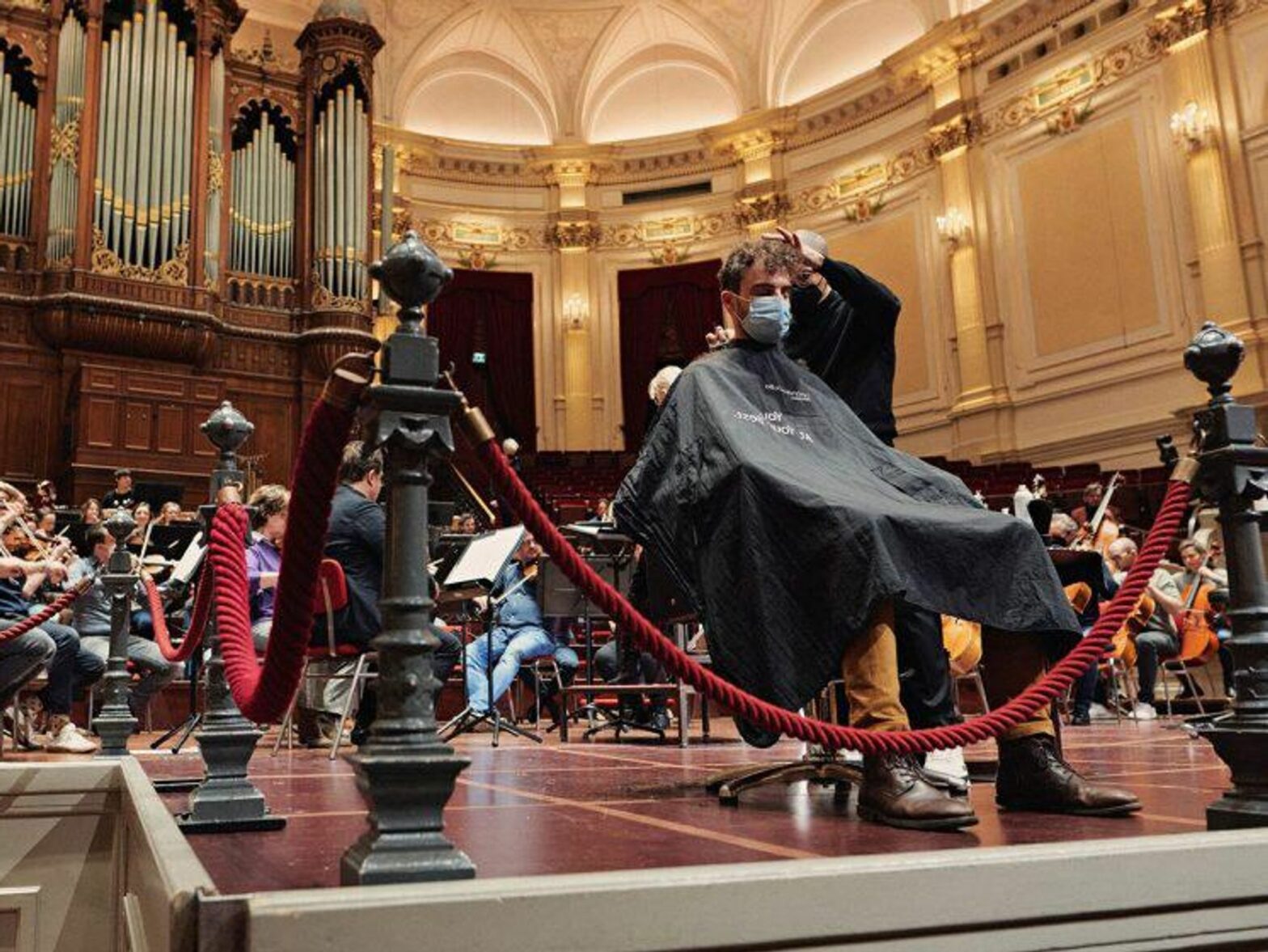 a man getting his hair cut while a symphonic band is playing in Amsterdam's concert hall