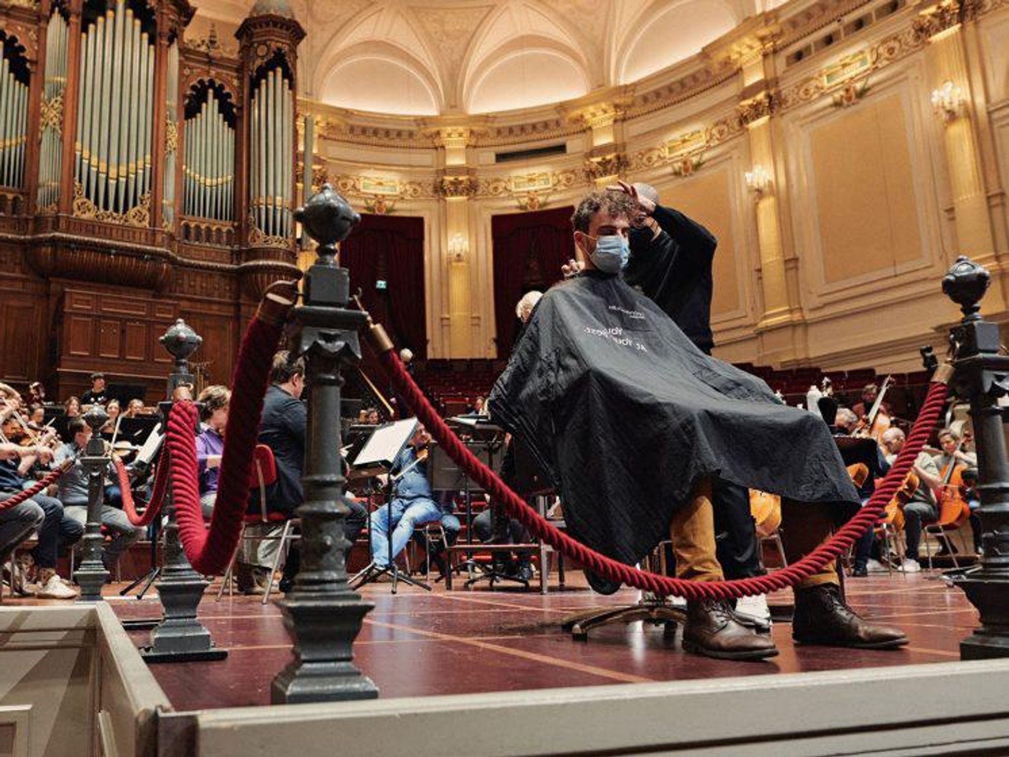 a man getting his hair cut while a symphonic band is playing in Amsterdam's concert hall