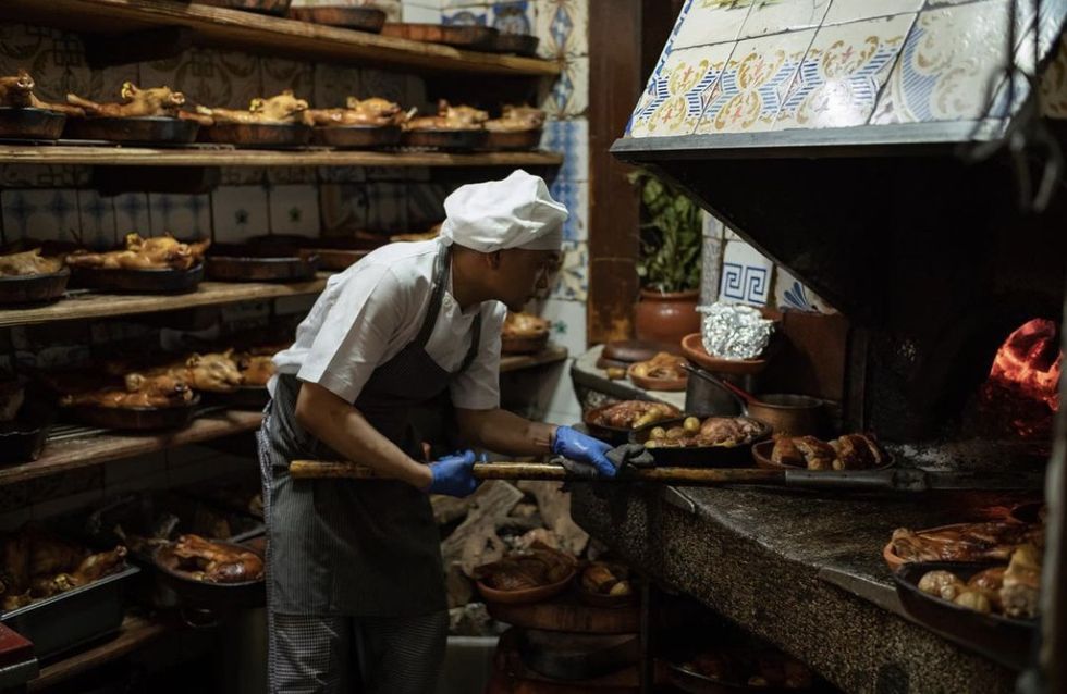 A man in a chefs hat tends the oven, with shelves of roast pig behind him