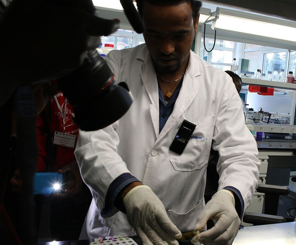 A man in a labcoat and latex gloves deals with samples in a lab, surrounded by reporters with cameras