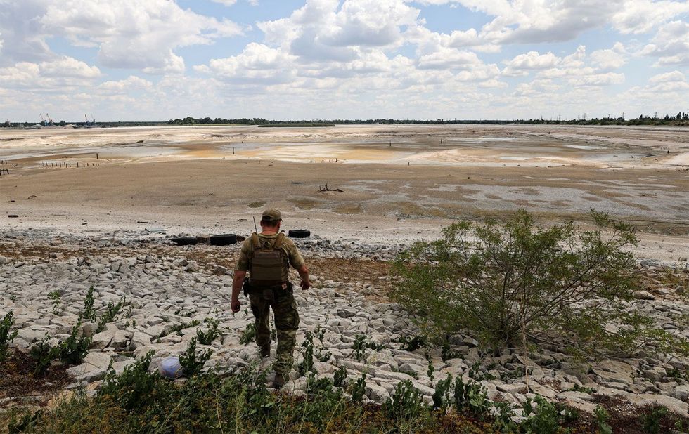 A man in fatigues walks down the stony bank to the dry reservoir bed.