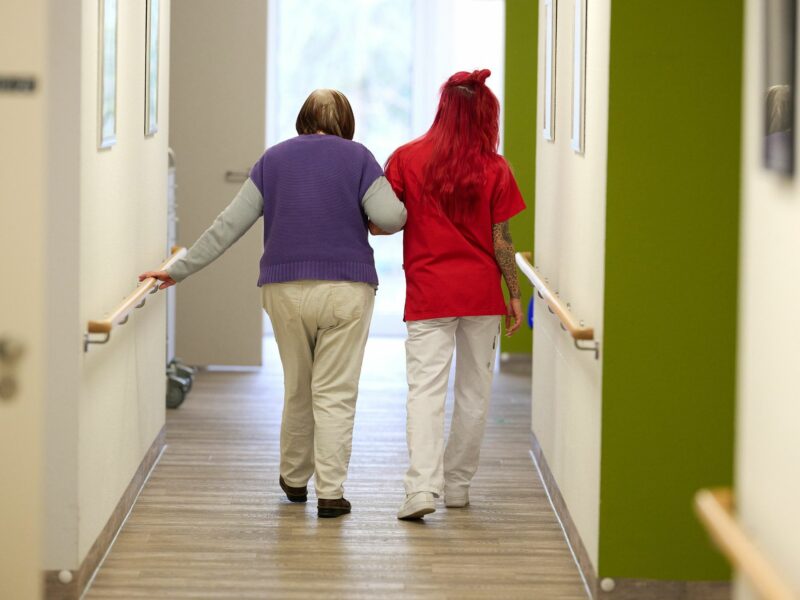 A nurse in a red shirt and an elderly patient in a purple sweater are seen from behind down the hall.