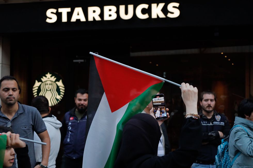 A Palestine flag is seen infront of the Starbucks logo and name as a woman is recording. There is a large crowd.