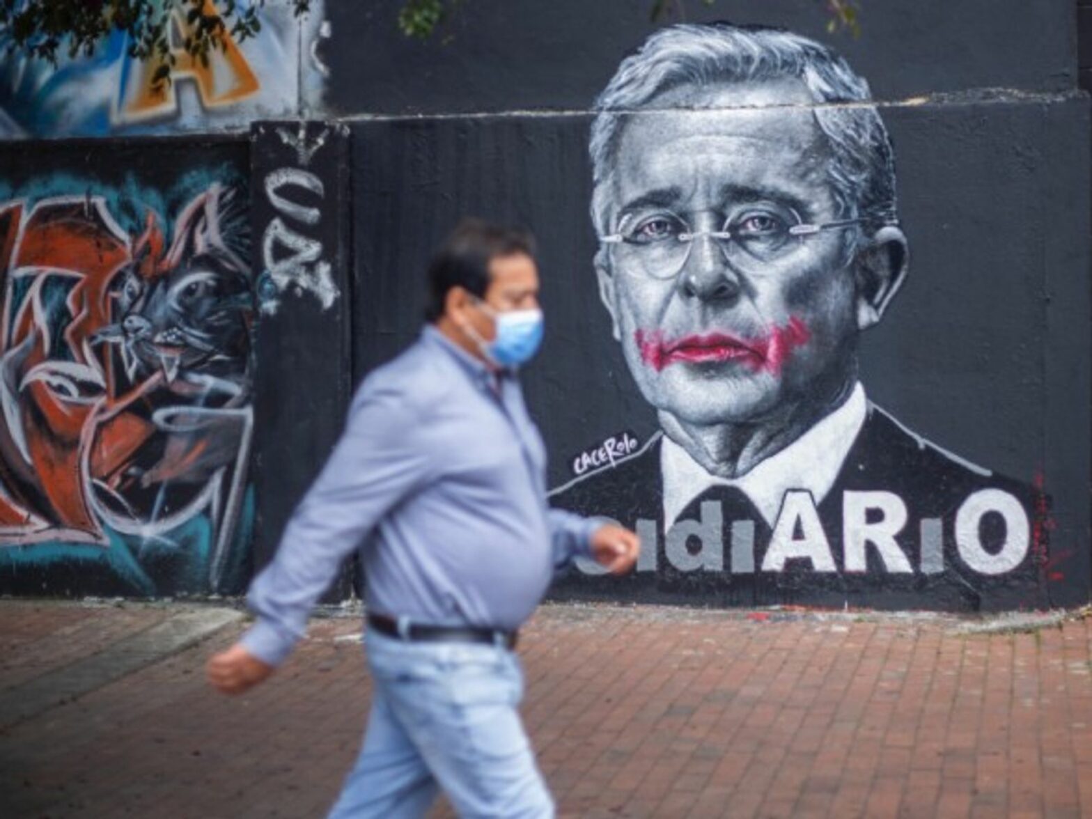 A person with a mask walking next to the image of former president Alvaro Uribe Velez in Bogota