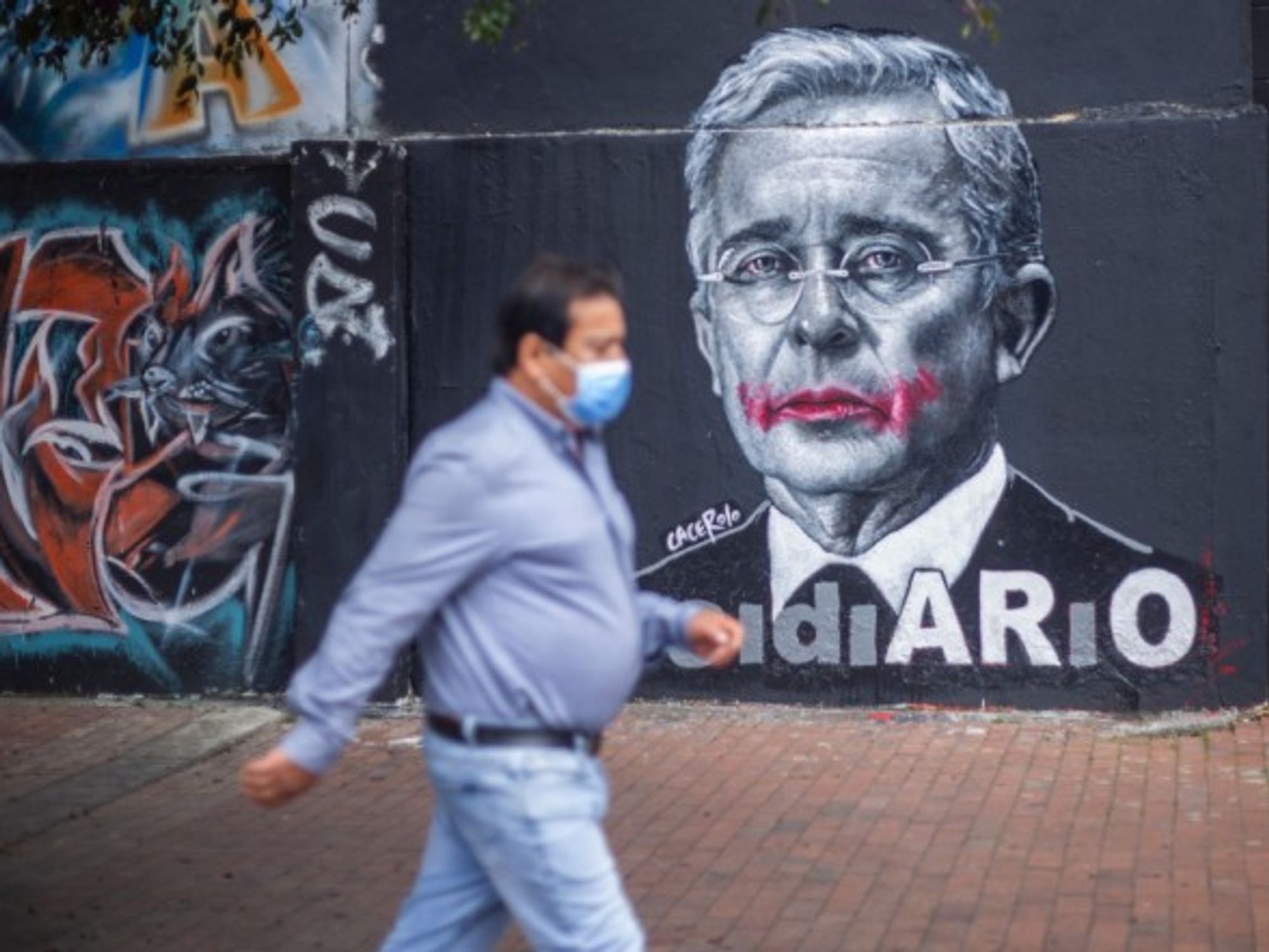 A person with a mask walking next to the image of former president Alvaro Uribe Velez in Bogota