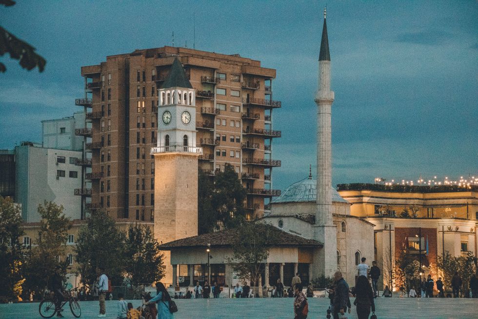 A photo of a square by night in Tirana, Albania. The square is lit up by surrounding buildings, including a clock tower. There are people moving about the space.