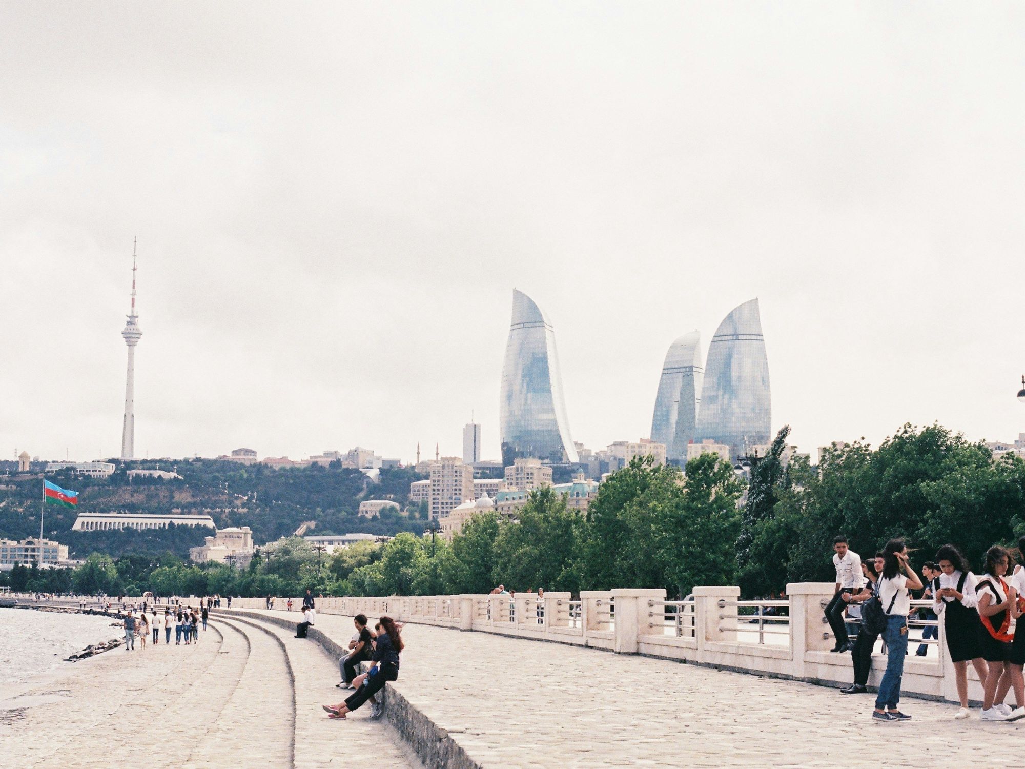 A photo of People enjoy the weather on the Baku boulevard. The iconic Flame Towers can be seen in the distance as well as the TV tower and a large flag.