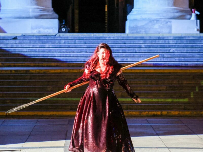 A photograph of Lucia Lucas singing with a lance, dressed in a black gown.