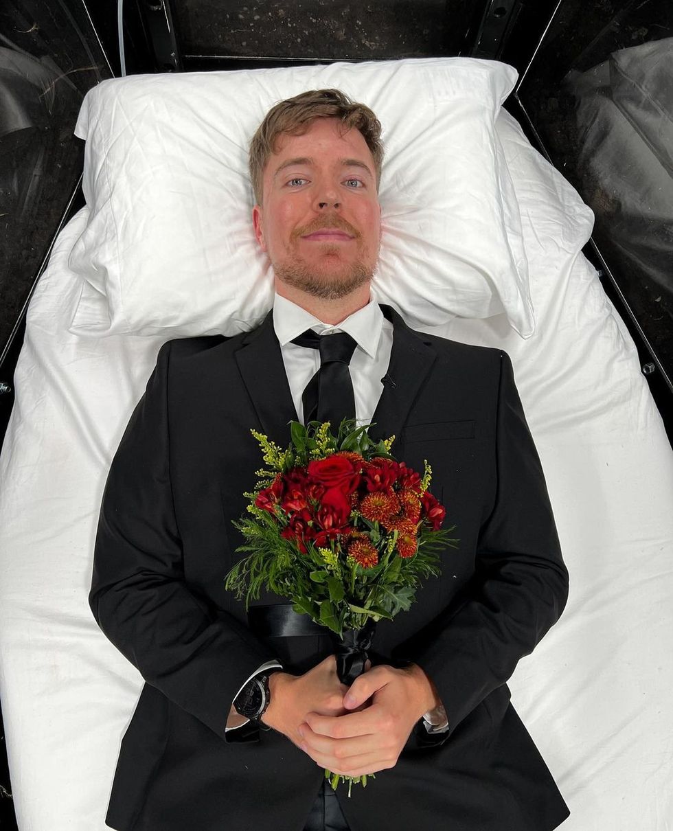 A photograph of mr beast in a suit holding flowers as he lies in a coffin