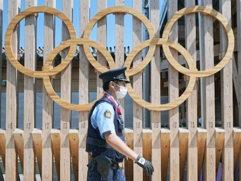 A police officer wearing a mask walks past Olympic rings at the entrance to Olympic Village