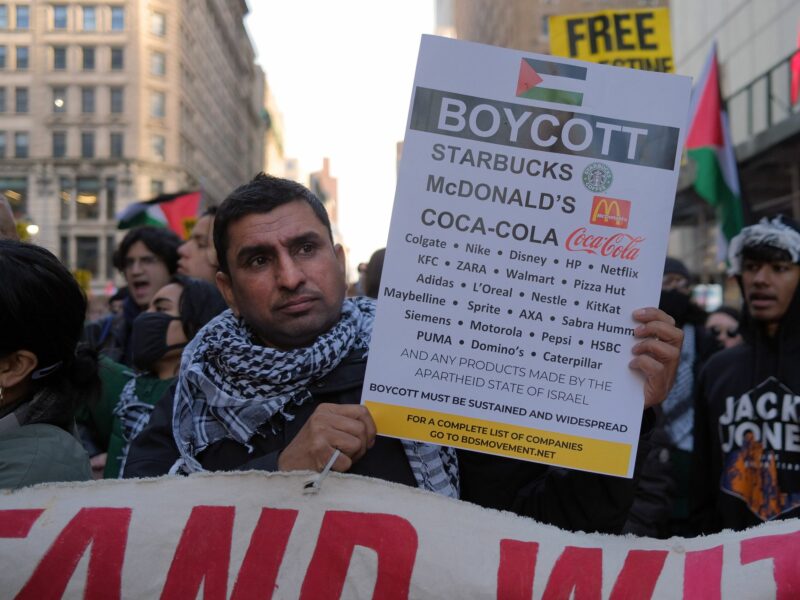 A protestor wearing the Palestinan keffiyah holding up a BDS sign with a list of companies to boycott.