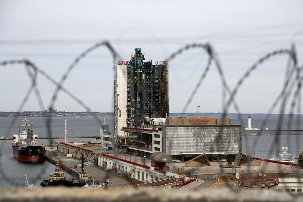 A small ship is docked at a port, next to a large and heavily damaged building with broken glass and entire parts missing.