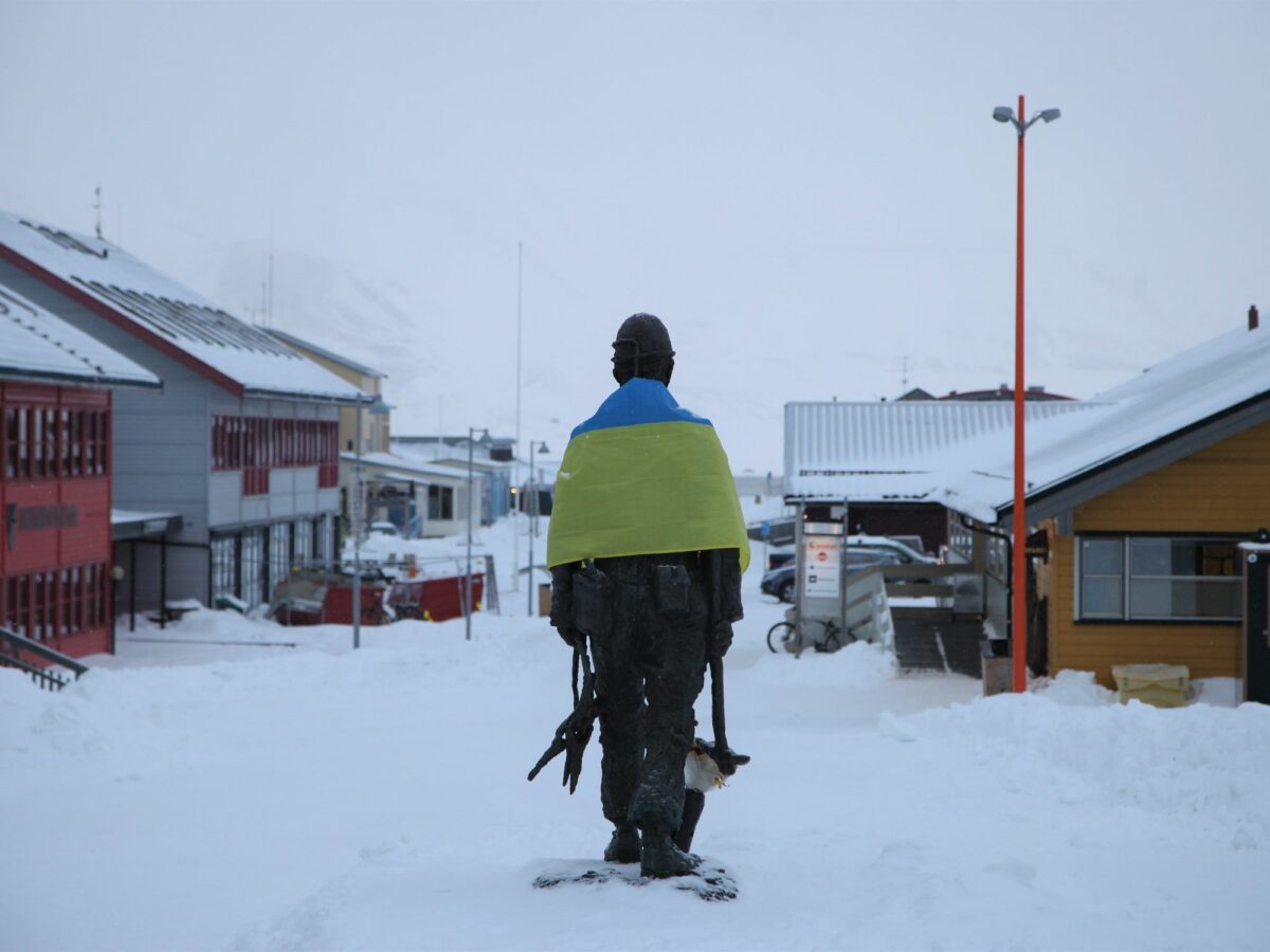 A statue of a coal miner stands in the center of the photos with houses surronding it, draped around their shoudler is a Ukrainian flag. The environment is snowy and the sky is white from clouds.