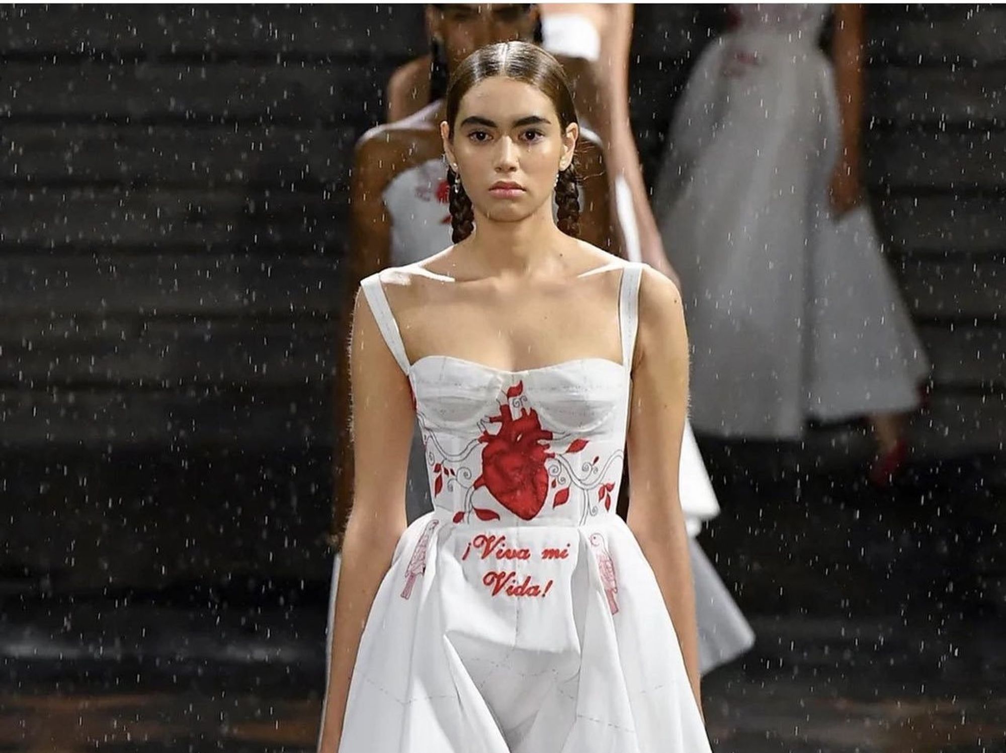 A woman in a white dress with red embroidery walks a catwalk in the rain
