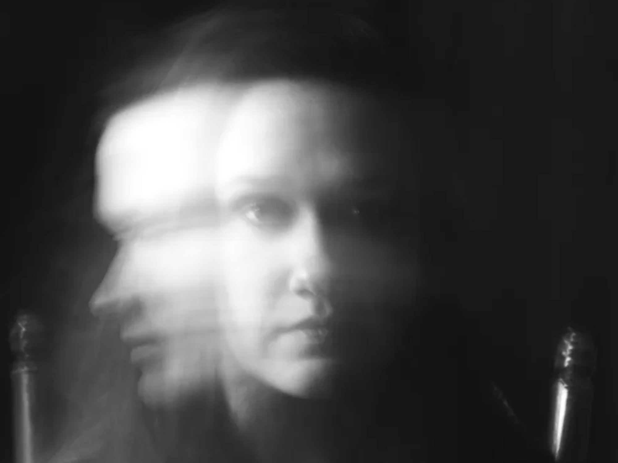 A woman in front of a black background moving her face quickly in the camera so that her face is not clearly captured.