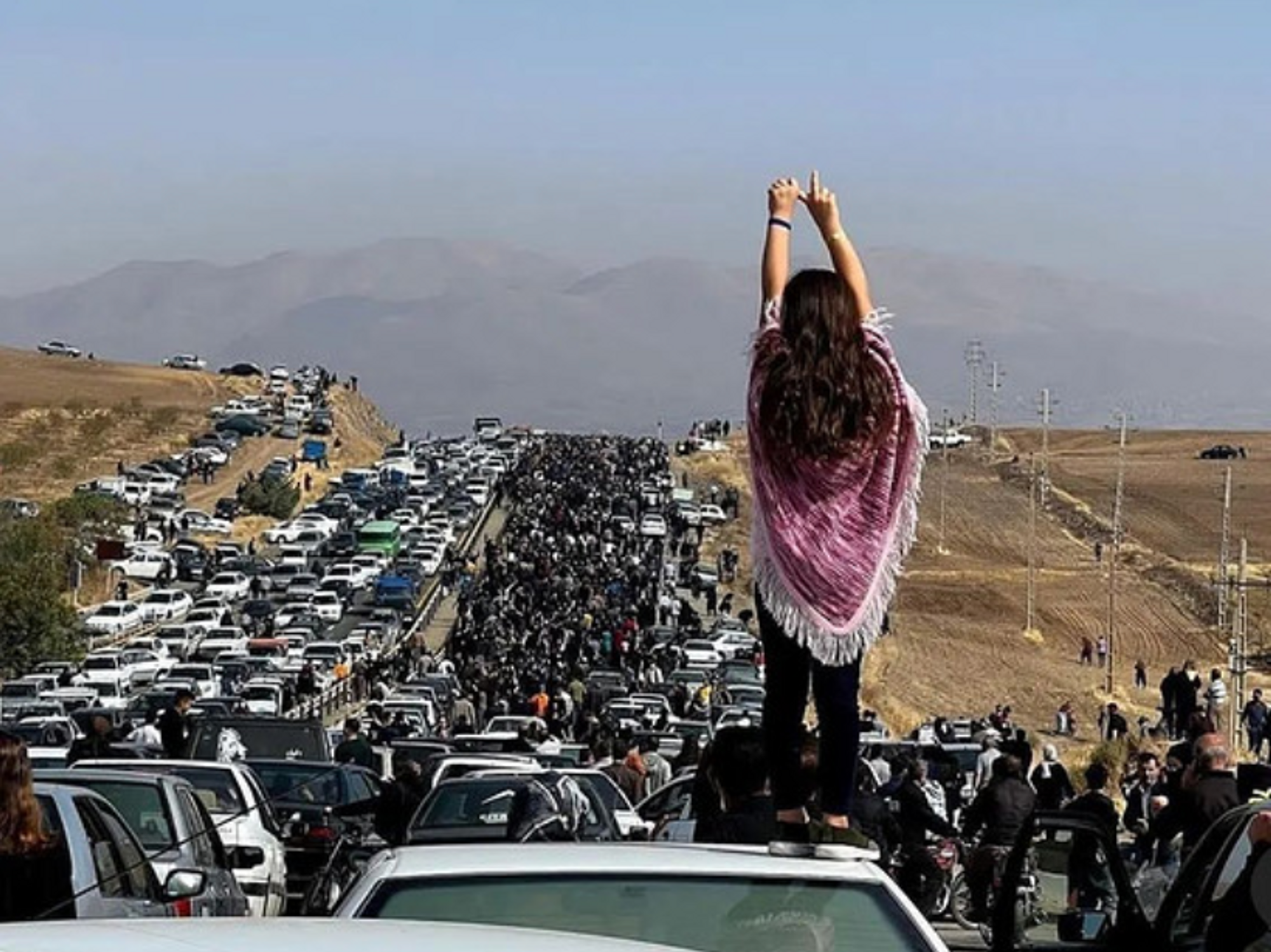 A woman stands on a car, in a road full of cars that have stopped. The young woman is raising her arms and is surrounded by protestors walking on the road.