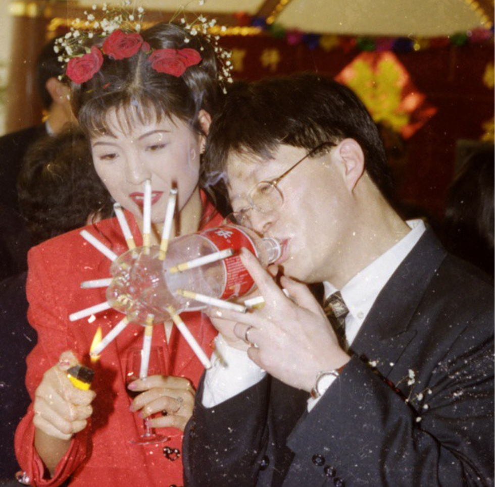 A women helps light a cigarette contraption for a man, in which many cigarettes are lit inside holes on the outside of a plastic soda bottle as the man inhales from the top.