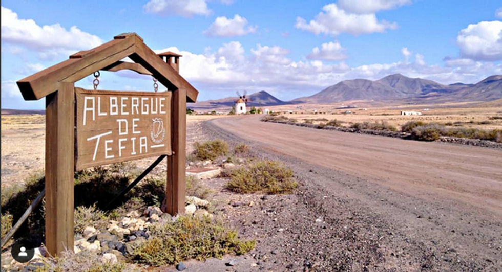 A wooden sign advertises the Tefía Hostel besides a dirt road, a mill and mountains in the distance.