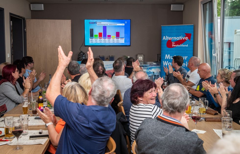 Adults at a meeting, sitting at tables, all in the process of clapping, some while looking at a screen at the front of the room