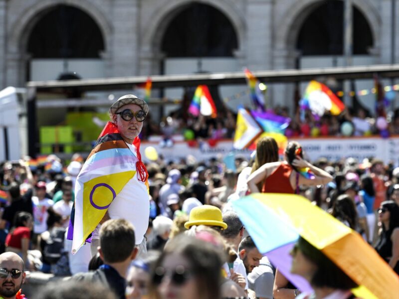 Amongst the Pride crowd, an older person in large sunglasses wears the updated progress pride flag over their shoulders