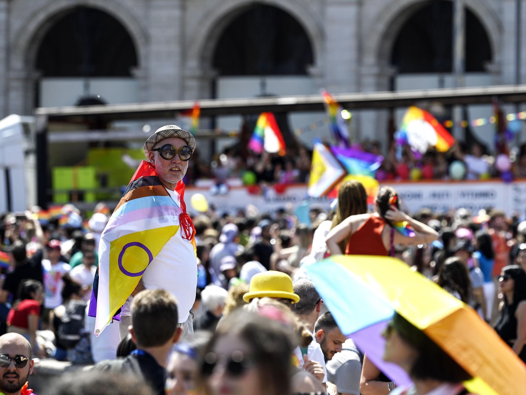 Amongst the Pride crowd, an older person in large sunglasses wears the updated progress pride flag over their shoulders