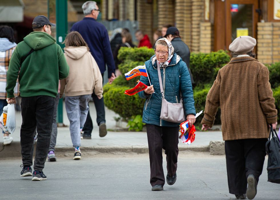 An elderly woman crosses the street carrying multiple flags